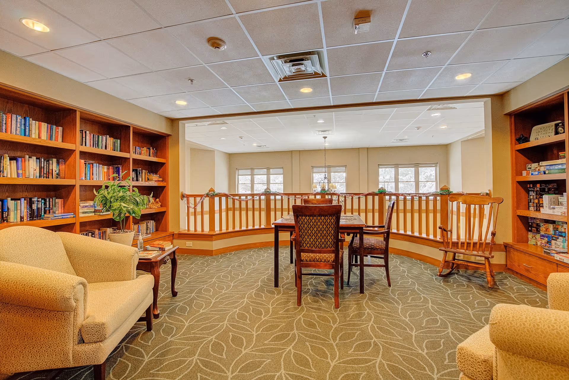 A cozy common area with bookshelves filled with books and board games on either side. There are two beige armchairs, a wooden table with four chairs in the center, and a wooden rocking chair to the right. The room has a patterned carpet and large windows letting in natural light.