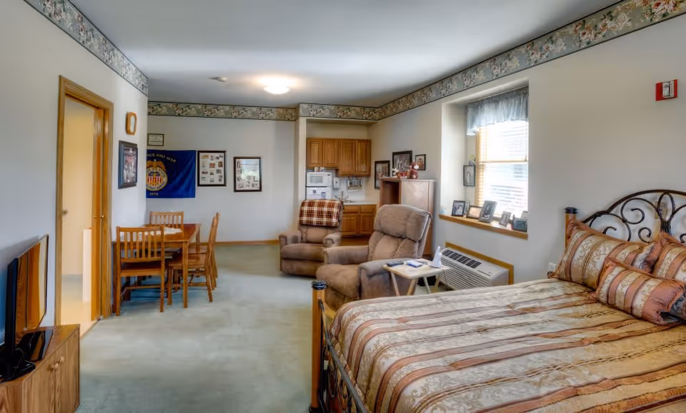 A senior living facility room featuring a bed with patterned bedding, two recliner chairs with a small table between them, a dining table with four chairs, a TV on a wooden stand, and a kitchenette with wooden cabinets and a microwave. The room has light-colored walls with floral wallpaper border near the ceiling, a window with blinds and a valance, and framed pictures on the walls.