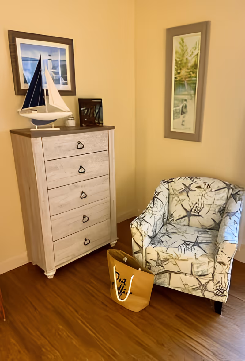 Corner of a living room with a white tall dresser topped by a model sailboat and framed art, next to a patterned armchair and a tote bag on a wood floor.