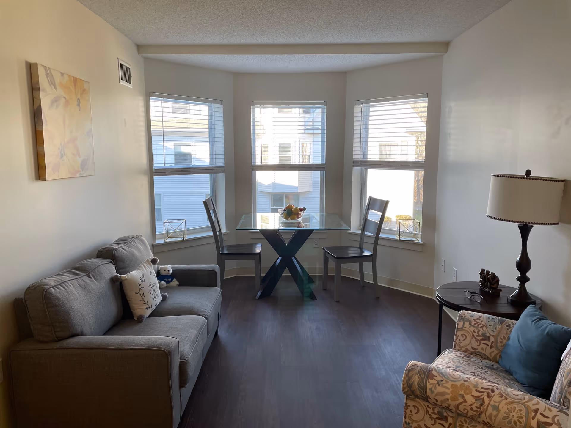 A cozy living room area with a gray sofa on the left, a patterned armchair with a blue pillow on the right, and a small round side table with a lamp and decorative items. In front of three large windows with blinds is a glass-top dining table with two chairs. The room has wooden flooring and light-colored walls with a floral painting hanging above the sofa.