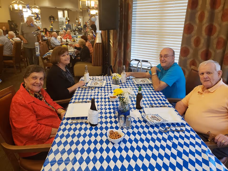 Four elderly people sitting around a table covered with a blue and white checkered tablecloth in a dining room. The table has plates, napkins, bottles, and small bowls of snacks. In the background, more elderly people are seated at other tables, and the room has warm lighting and large windows with curtains.