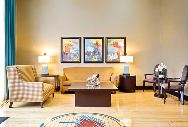 A well-lit living room with a beige sofa centered against a wall, flanked by two side tables each with a lamp. There is a patterned armchair on the left and two wooden chairs with a small round table on the right. Three colorful floral paintings hang above the sofa. The floor is light wood with a circular decorative design in the foreground.