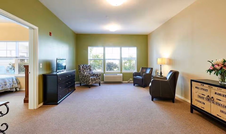 Spacious assisted living common room with chairs, a TV on a dresser, a lamp, large window, and a doorway into a bedroom.