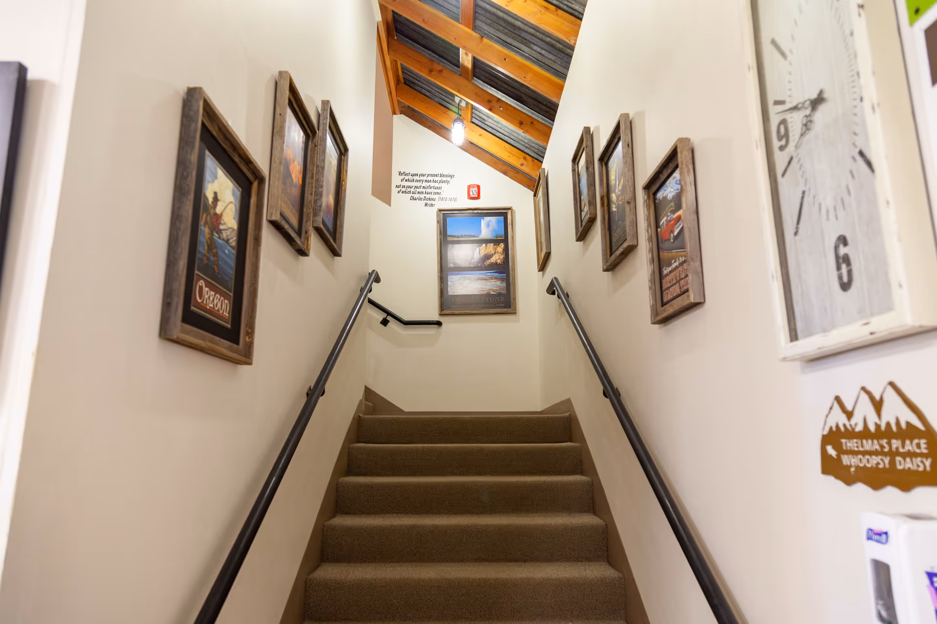 Carpeted staircase with black handrails on both sides, walls decorated with framed pictures and a large clock. The ceiling above the stairs has wooden beams and a hanging light fixture.