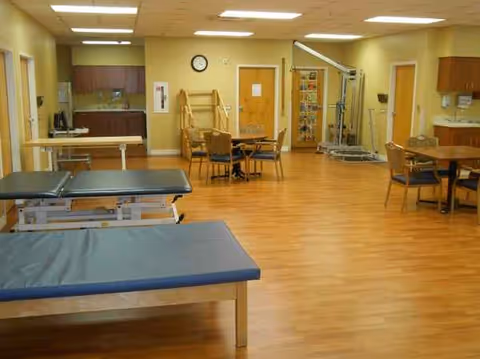Rehabilitation therapy room with treatment tables, chairs, and exercise equipment in a sunny facility