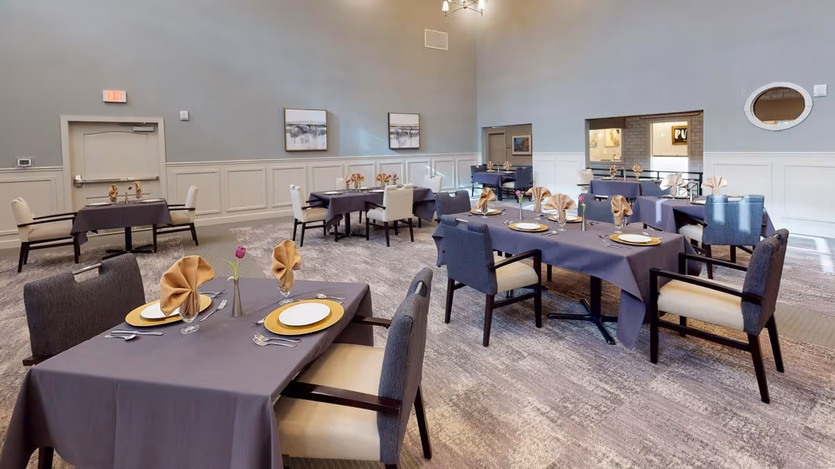 A dining room with several tables covered in dark tablecloths, each set with plates, gold chargers, folded napkins, and glassware. The room has light gray walls with white wainscoting, carpeted floors, and framed artwork on the walls. Chairs with dark frames and light cushions surround the tables.
