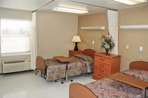 Two hospital-style beds with floral bedding, a dresser, lamp, and window in a shared room.