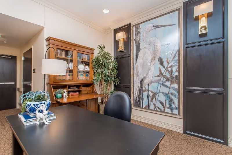An interior office or study area with a black desk and black chair in the foreground. On the desk are decorative items including a blue and white ceramic pot with a plant and a small white animal figurine. Behind the desk is a wooden cabinet with glass doors containing books and decorative objects. A tall green plant is placed next to the cabinet. On the wall is a large framed artwork of a white heron bird with two wall sconces on either side. The room has beige walls and carpeted floor.