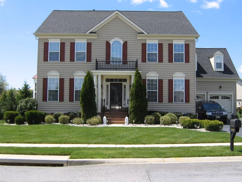 Front exterior view of a two-story beige house with red shutters, a black front door with sidelights, a small balcony above the entrance, neatly trimmed bushes and trees in the front yard, a driveway with a black SUV parked, and a mailbox near the street.