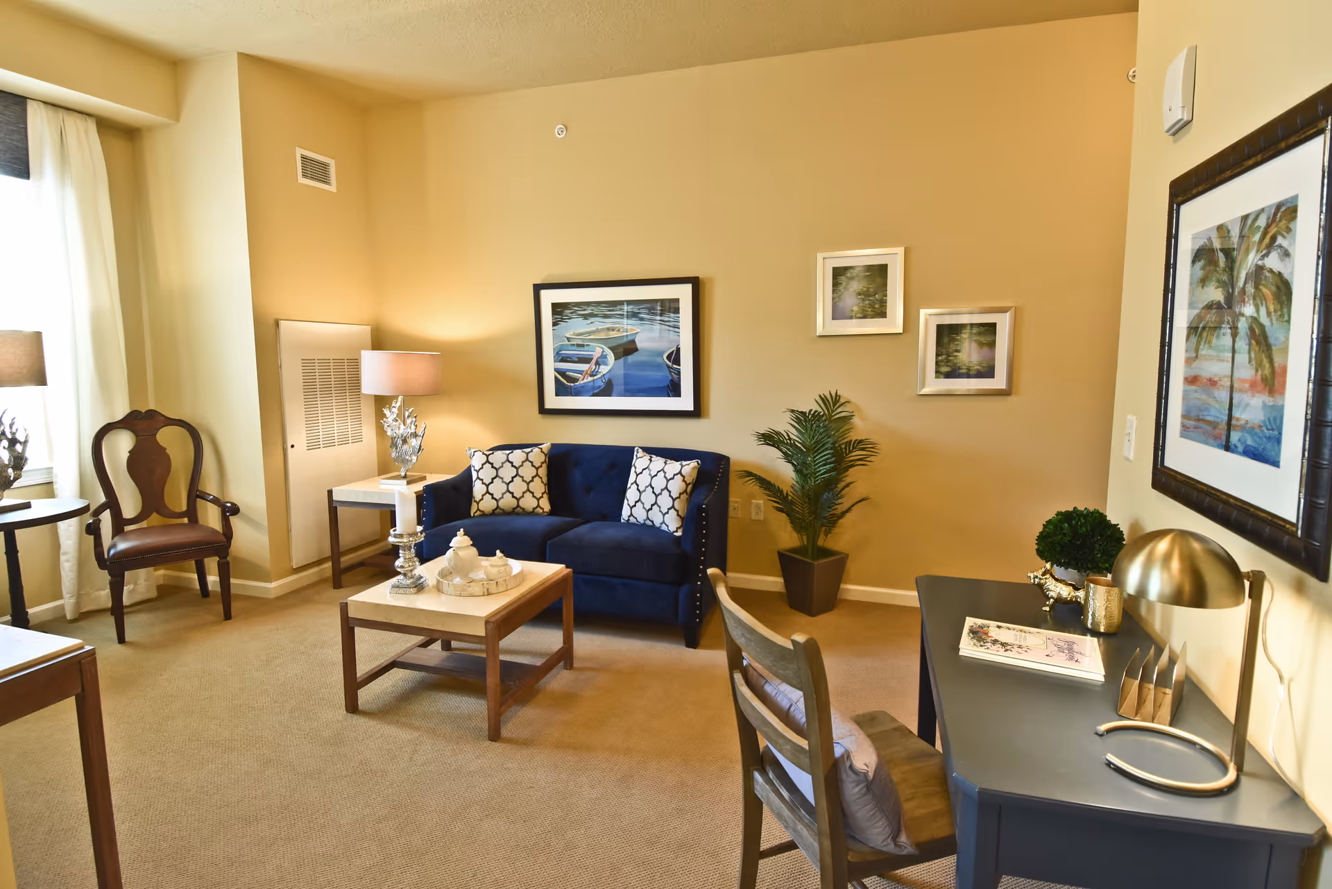 Bright furnished living room with a navy sofa, coffee table, desk, and framed artwork on beige walls.