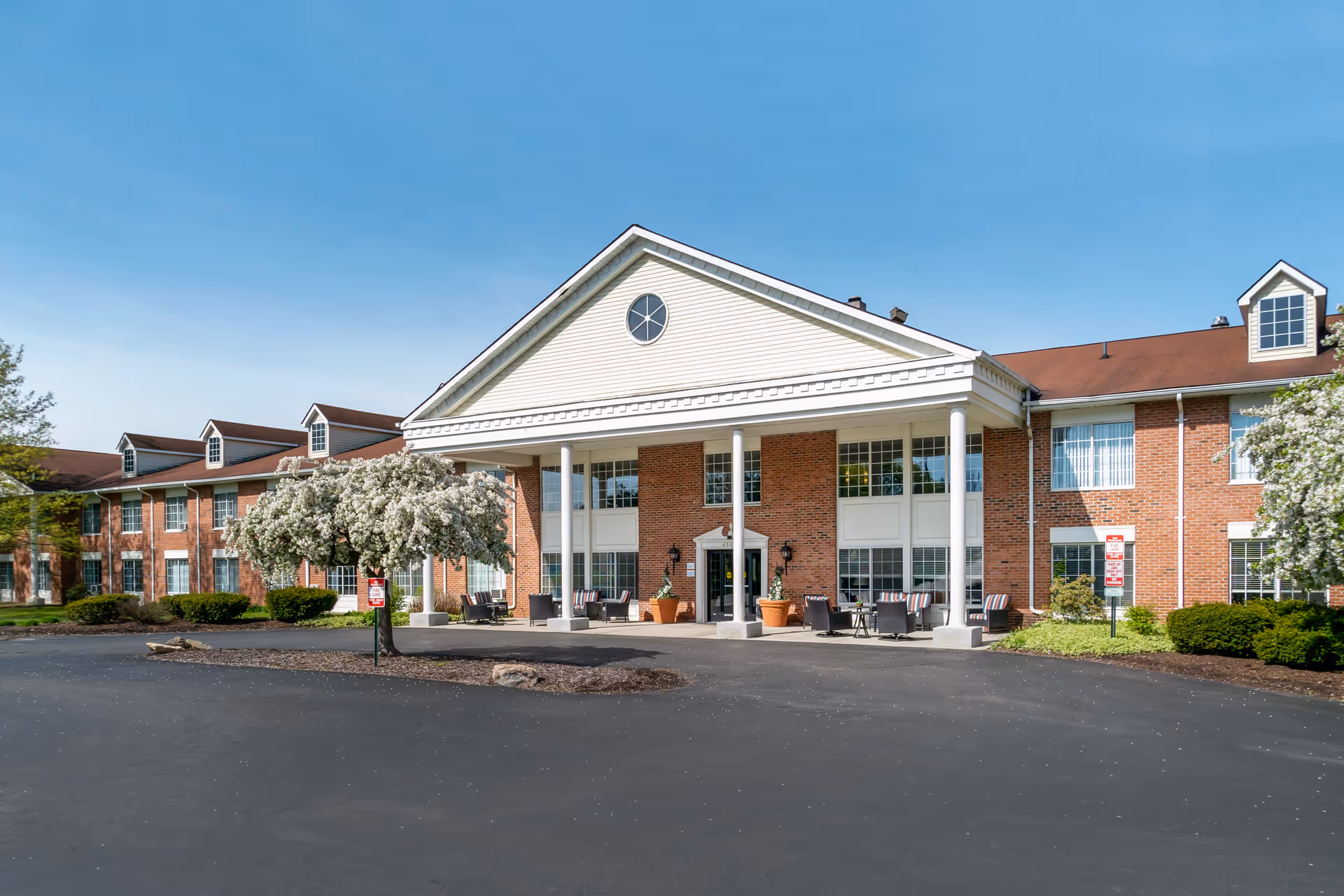 Front exterior view of a two-story brick building with white columns supporting a large triangular pediment above the entrance. There are several windows, outdoor seating areas with chairs and tables, and blooming trees on either side of the entrance. The sky is clear and blue.