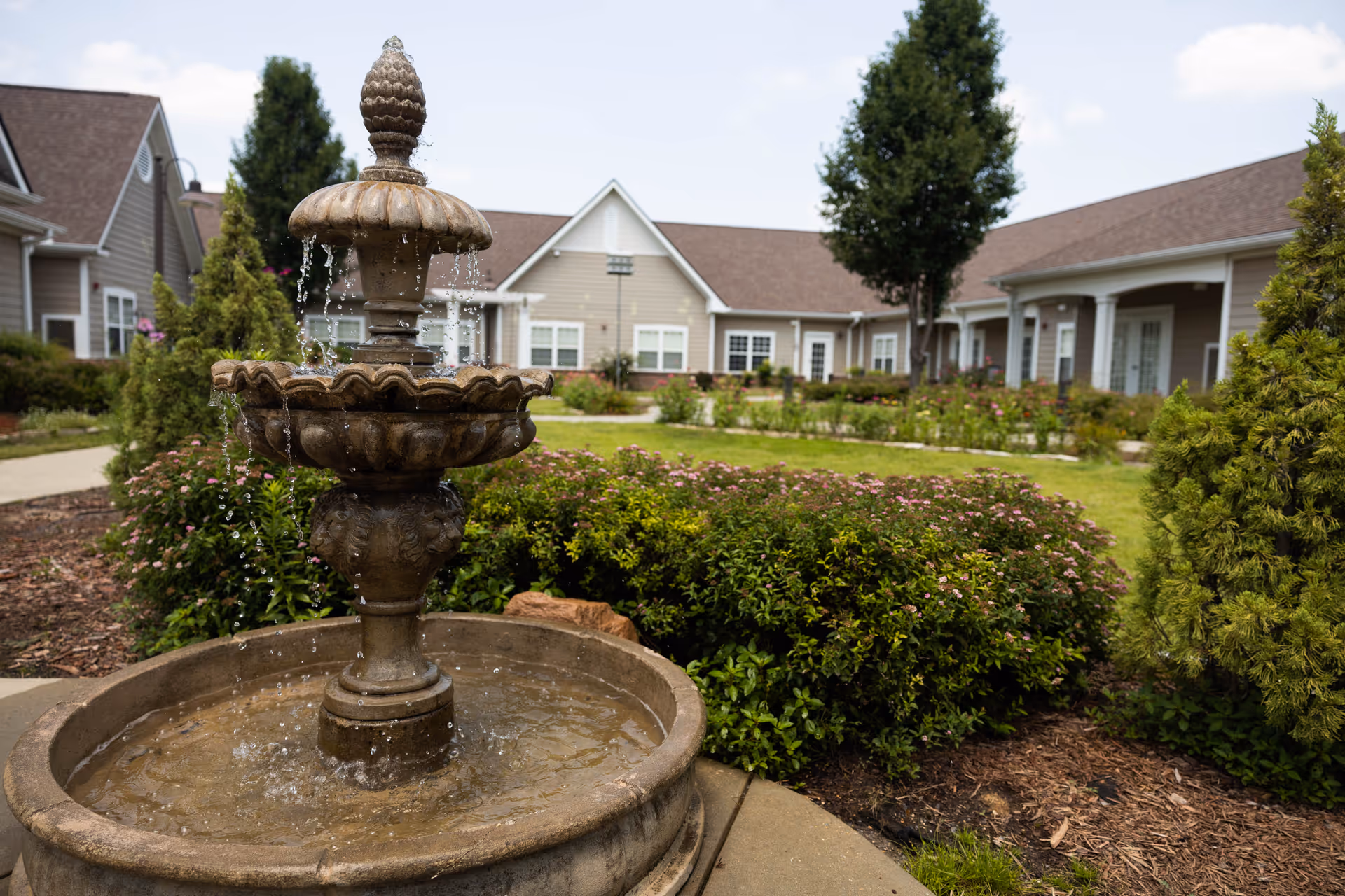 Outdoor courtyard area of a senior living facility with a decorative stone water fountain in the foreground, surrounded by green shrubs and flowering plants, with beige buildings and trees in the background under a partly cloudy sky.
