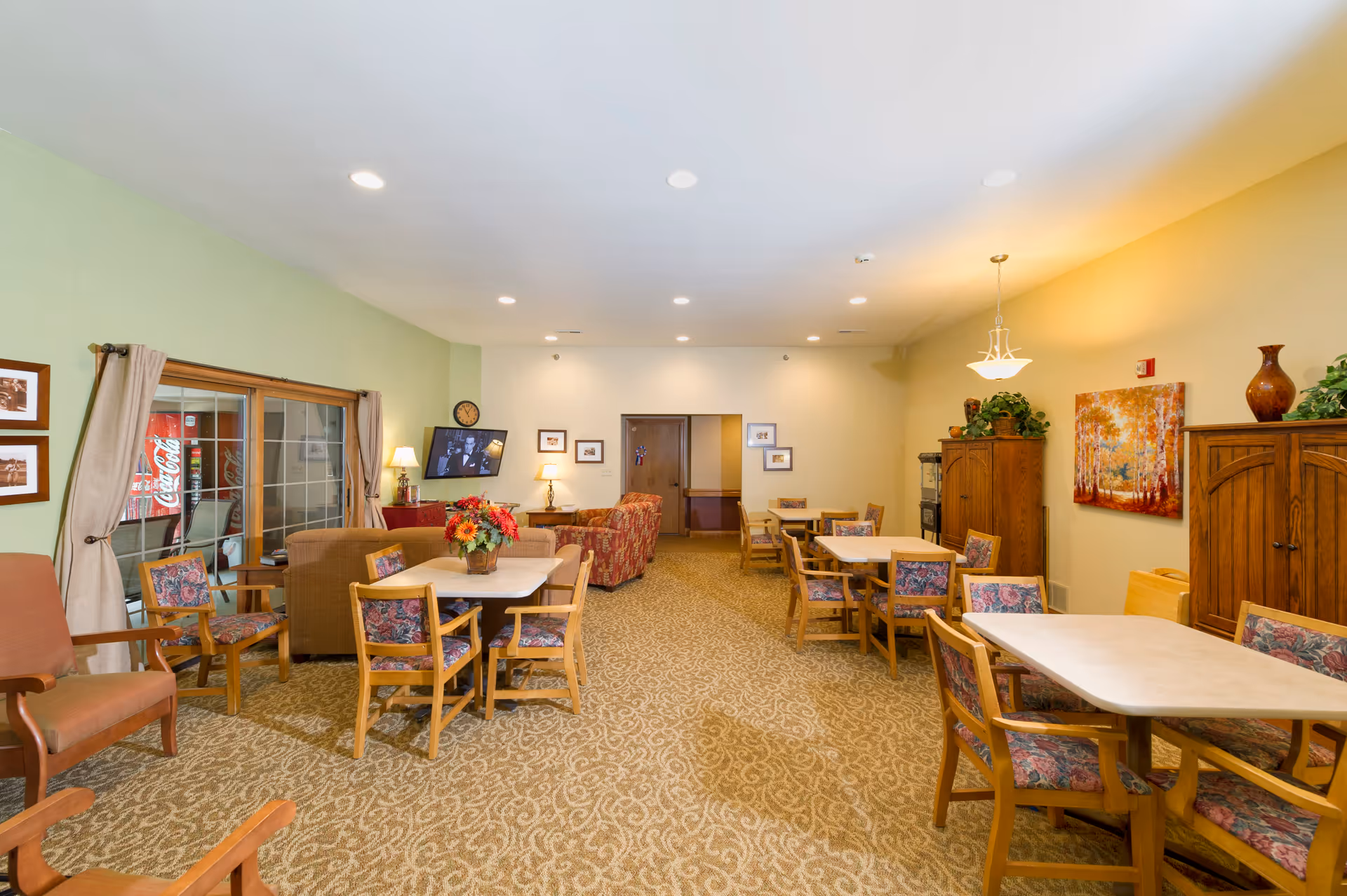 A well-lit common area in a senior living facility featuring multiple tables with floral upholstered chairs, a few armchairs, and a sofa. The walls are painted in light green and beige tones, decorated with framed pictures and a colorful painting. A flat-screen TV is mounted on the wall, and there are plants and decorative vases on wooden cabinets. A vending machine is visible through a glass door on the left side.