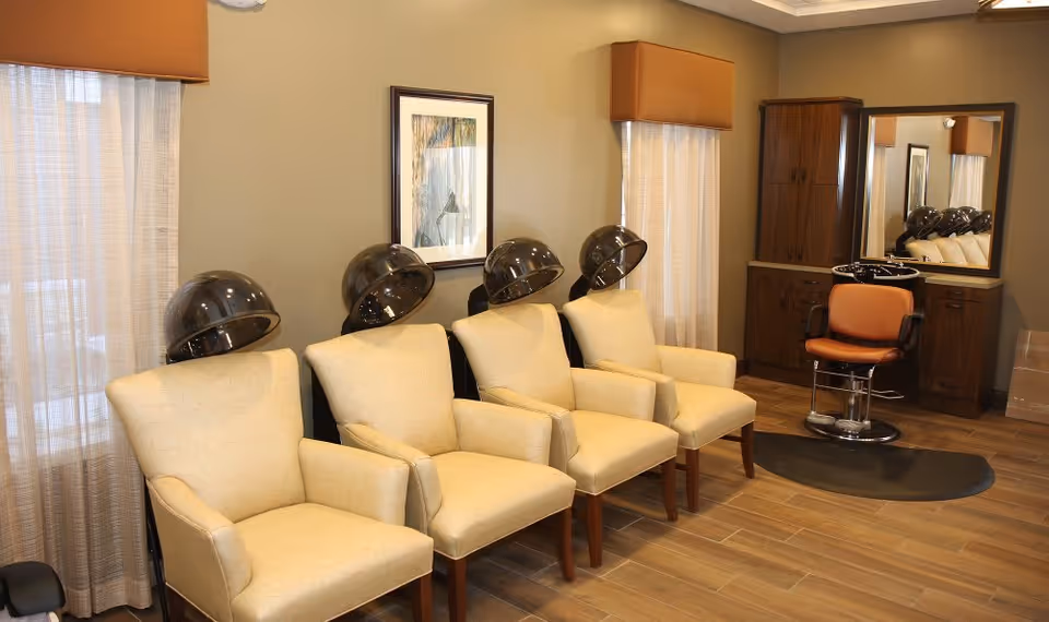 Interior view of a hair salon area in an assisted living facility featuring four beige salon chairs with hair dryers above them, a single orange salon chair in front of a mirror and wooden cabinetry, with light brown walls and wood flooring.