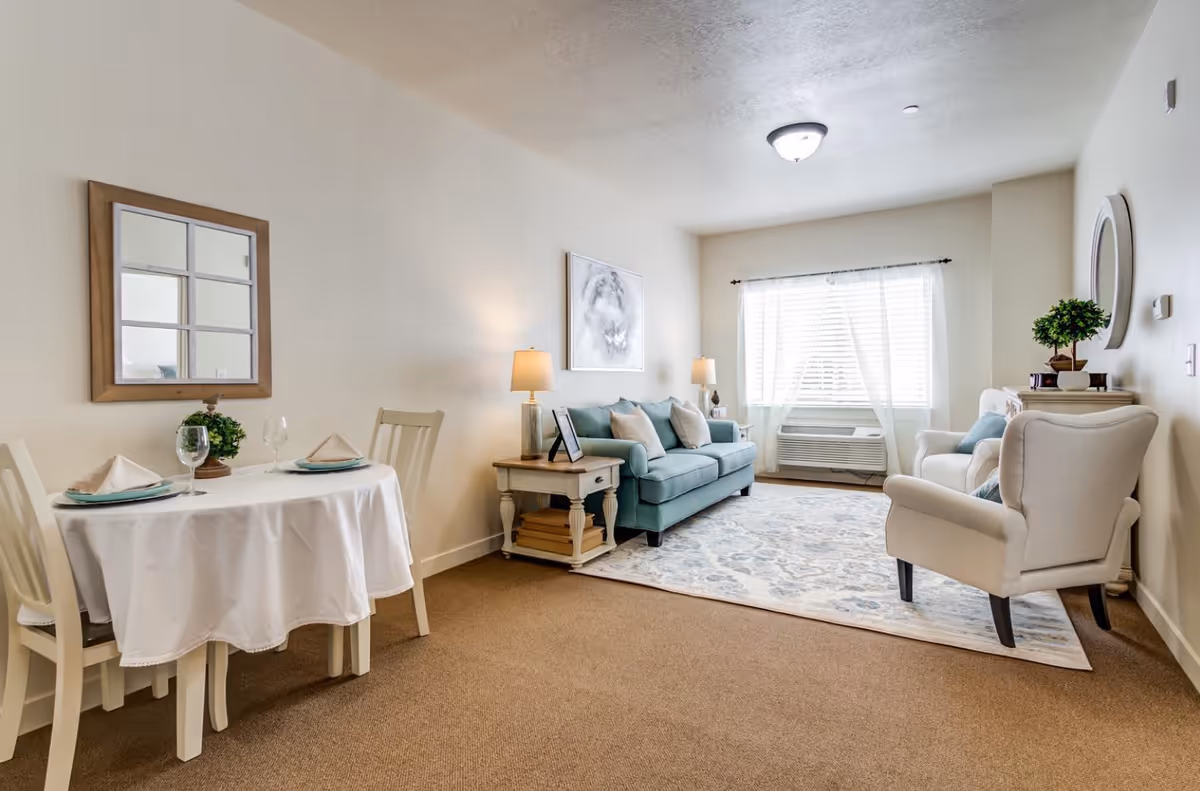 A bright and cozy living room area with a light blue sofa, two white armchairs, a patterned rug, and a window with sheer white curtains. To the left, there is a small dining table covered with a white tablecloth, set with two place settings including plates, napkins, and wine glasses. The walls are light-colored, and there are two table lamps on side tables, a framed mirror, and a piece of artwork on the wall.