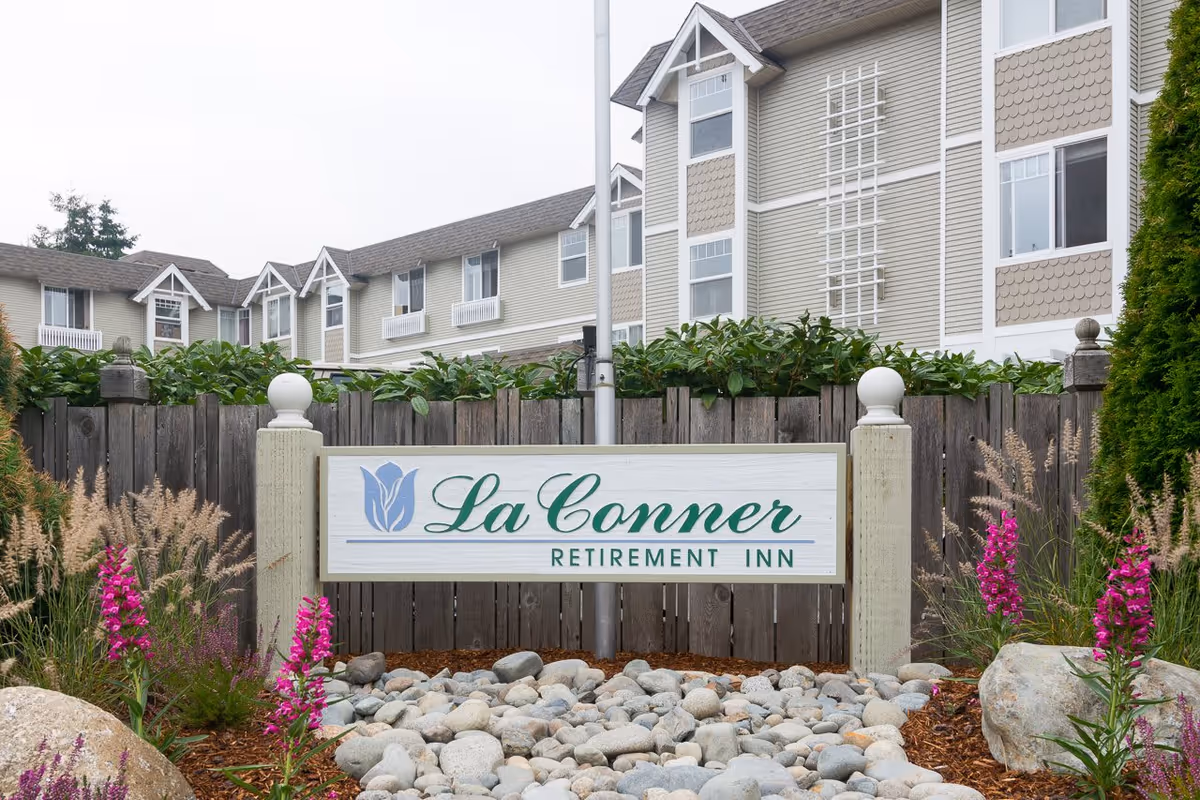 Outdoor view of La Conner Retirement Inn sign with a landscaped garden featuring rocks, purple flowers, and ornamental grasses in front of a multi-story residential building with beige siding and white trim.