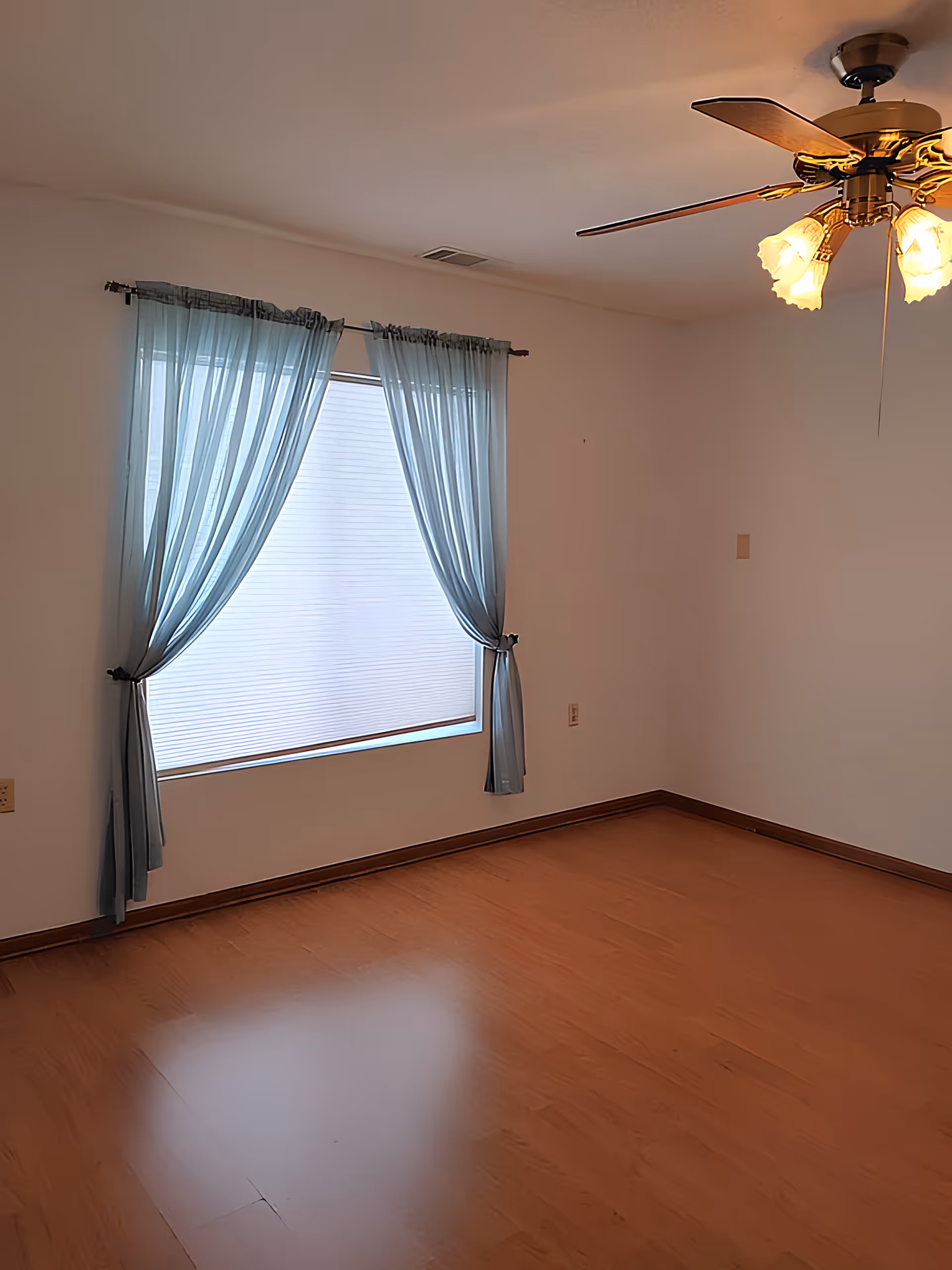 Empty room with hardwood floor, a curtained window, and a ceiling fan overhead.