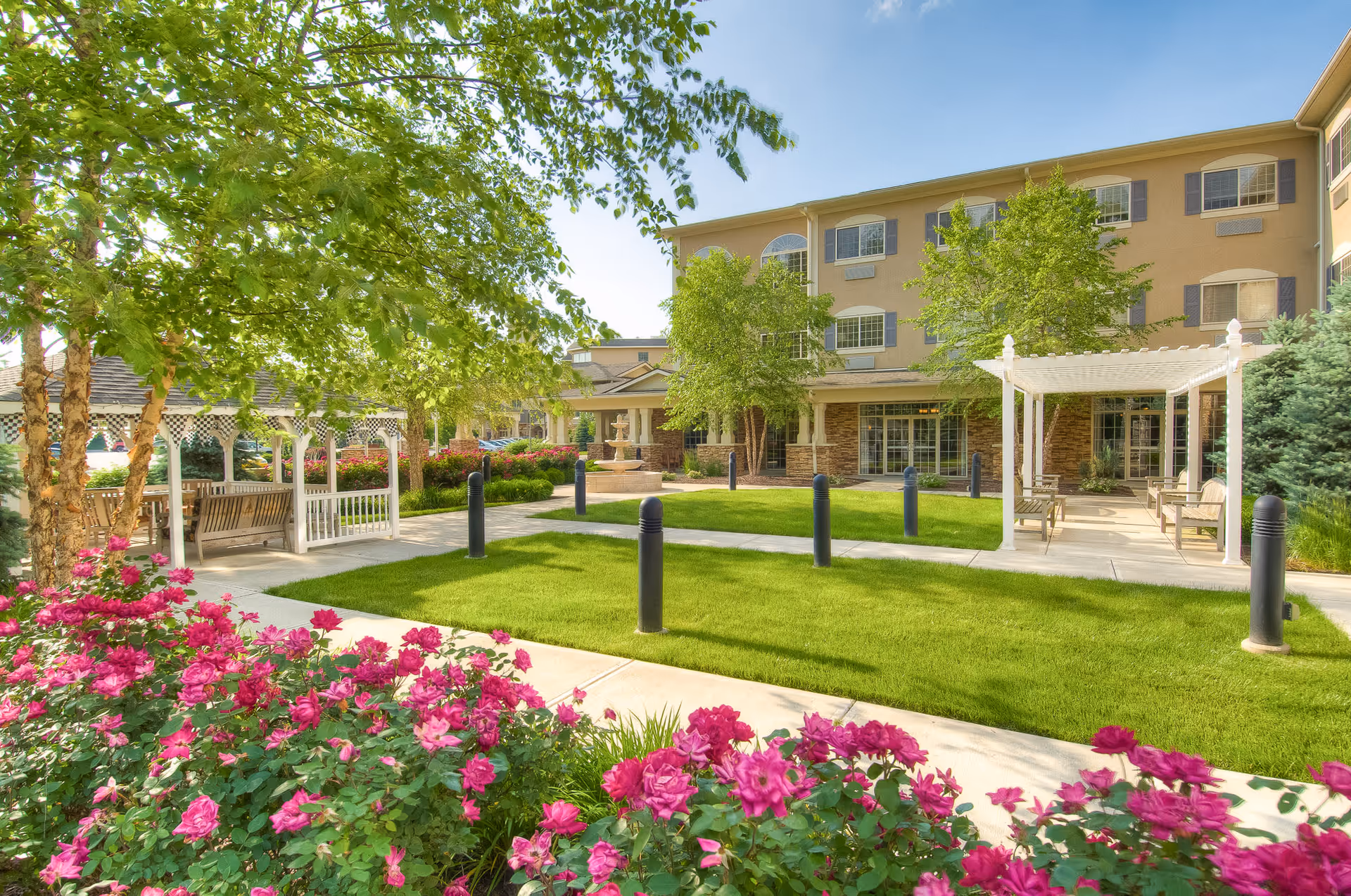 Sunlit courtyard with green lawn, pink flowers, pergolas and benches in front of a multi-story senior living building.