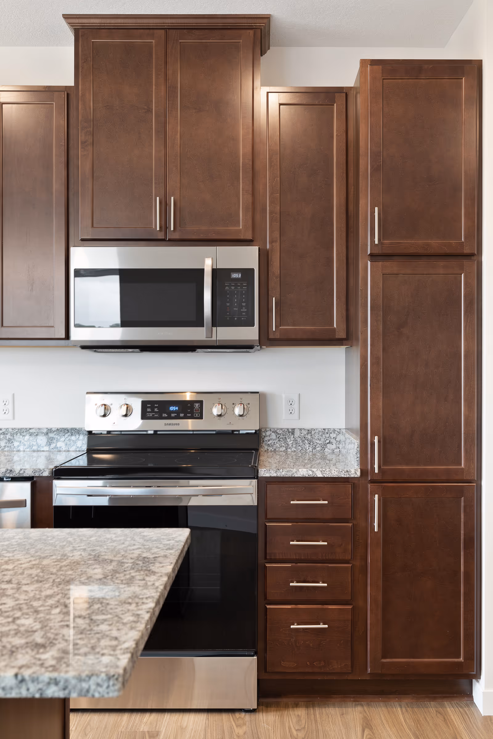 Modern kitchen with dark wooden cabinets, a stainless steel microwave mounted above a stainless steel electric stove, granite countertops, and a wooden floor.