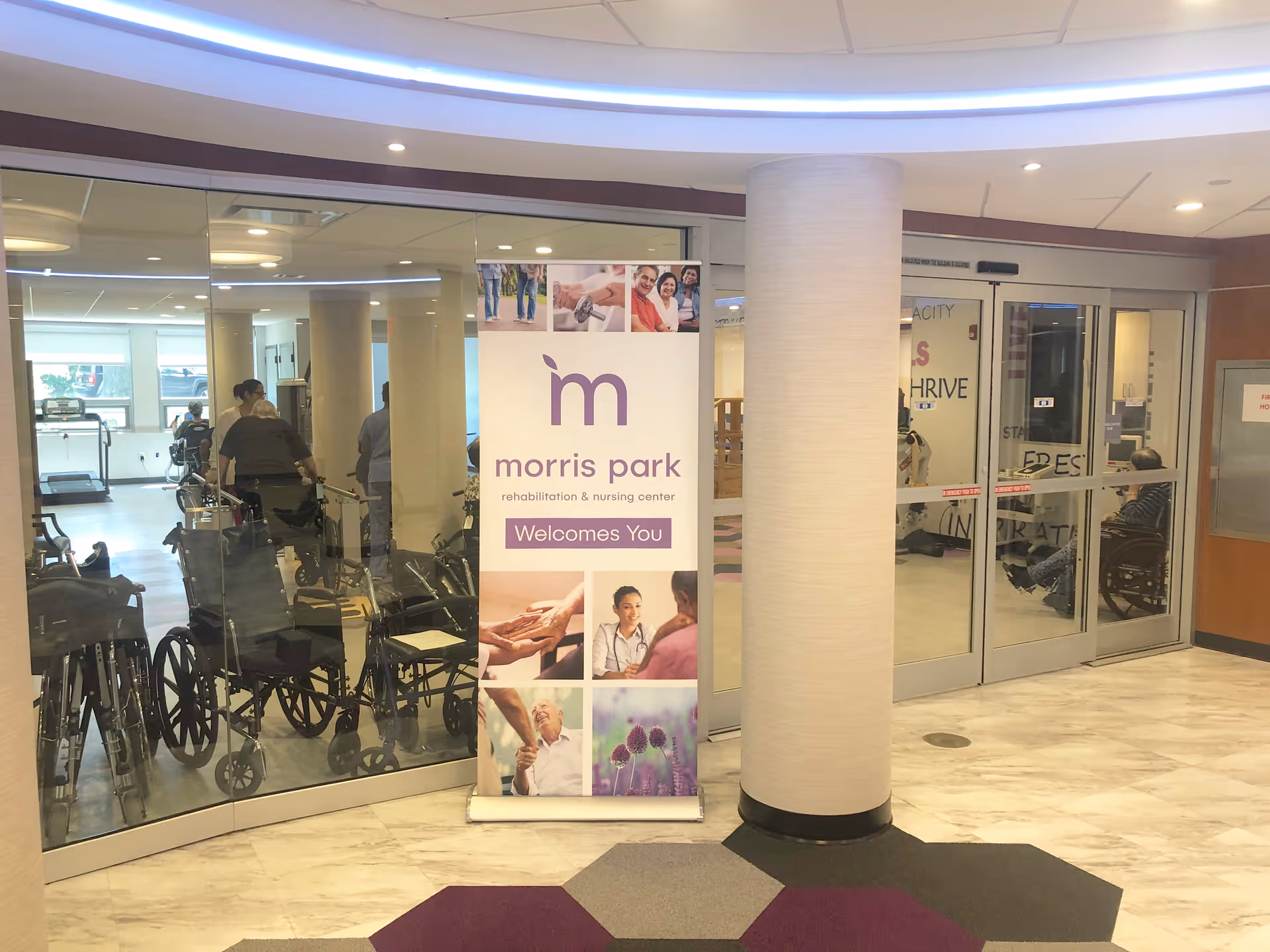 Interior view of Morris Park Rehabilitation & Nursing Center showing a glass wall with wheelchairs and people inside a room. A standing banner with the facility's name and welcoming message is placed near a round column. The floor has a patterned carpet and marble tiles.