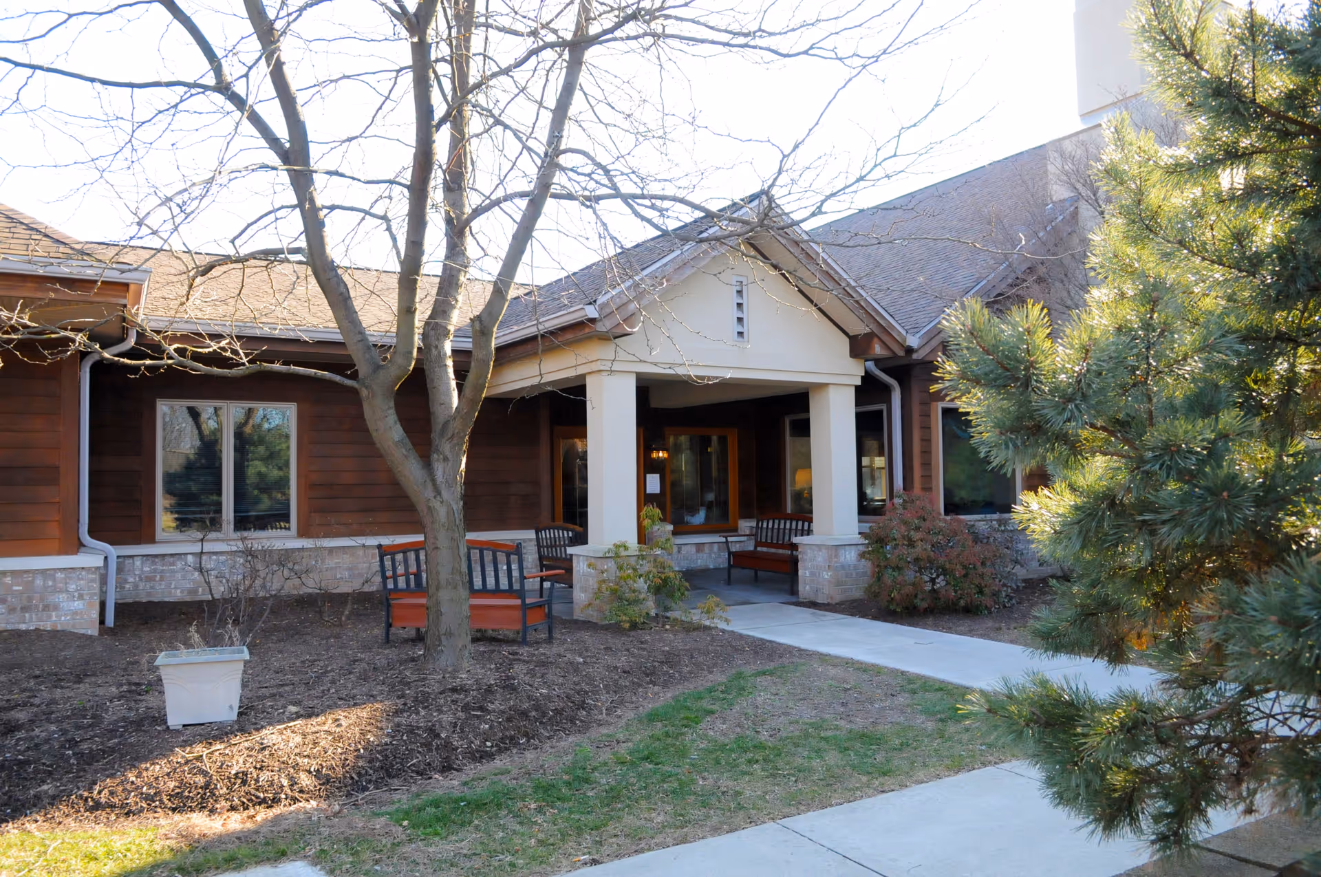 Front entrance of a one-story building with a covered porch, outdoor benches, a concrete walkway, and trees.