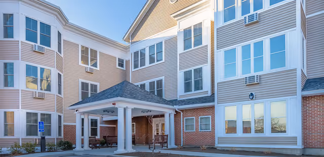 Exterior view of a multi-story senior living facility building with beige siding and red brick accents. The entrance features a covered drop-off area supported by white columns, with benches underneath. Several windows are visible, and the sky is clear and blue.