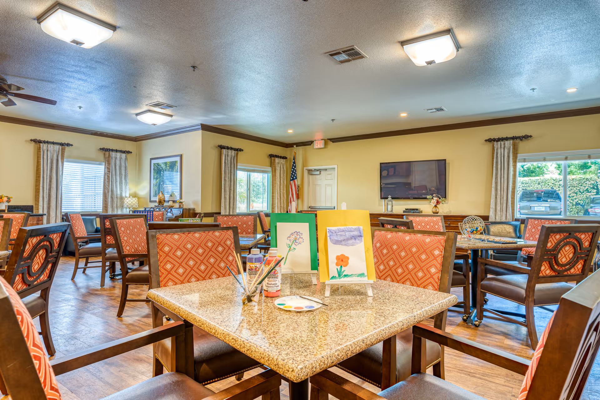 A well-lit common room with multiple tables and chairs featuring red patterned upholstery. On the closest table, there are two small easels displaying watercolor paintings of flowers, paint bottles, brushes, and a paint palette. The room has large windows with curtains, a television mounted on the wall, and an American flag near a door. The floor is wooden, and the ceiling has several light fixtures.