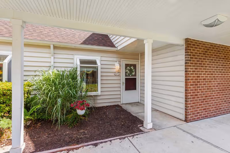 Covered entrance area to a residential unit with beige siding and a brick wall. There is a white door with a wreath, a window with a small awning, and a garden bed with green plants and a hanging pot of red flowers.