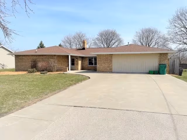 Single-story brick house with a large driveway, two-car garage, and a small front porch. The house is surrounded by a lawn with some bushes near the entrance and leafless trees in the background under a clear blue sky.