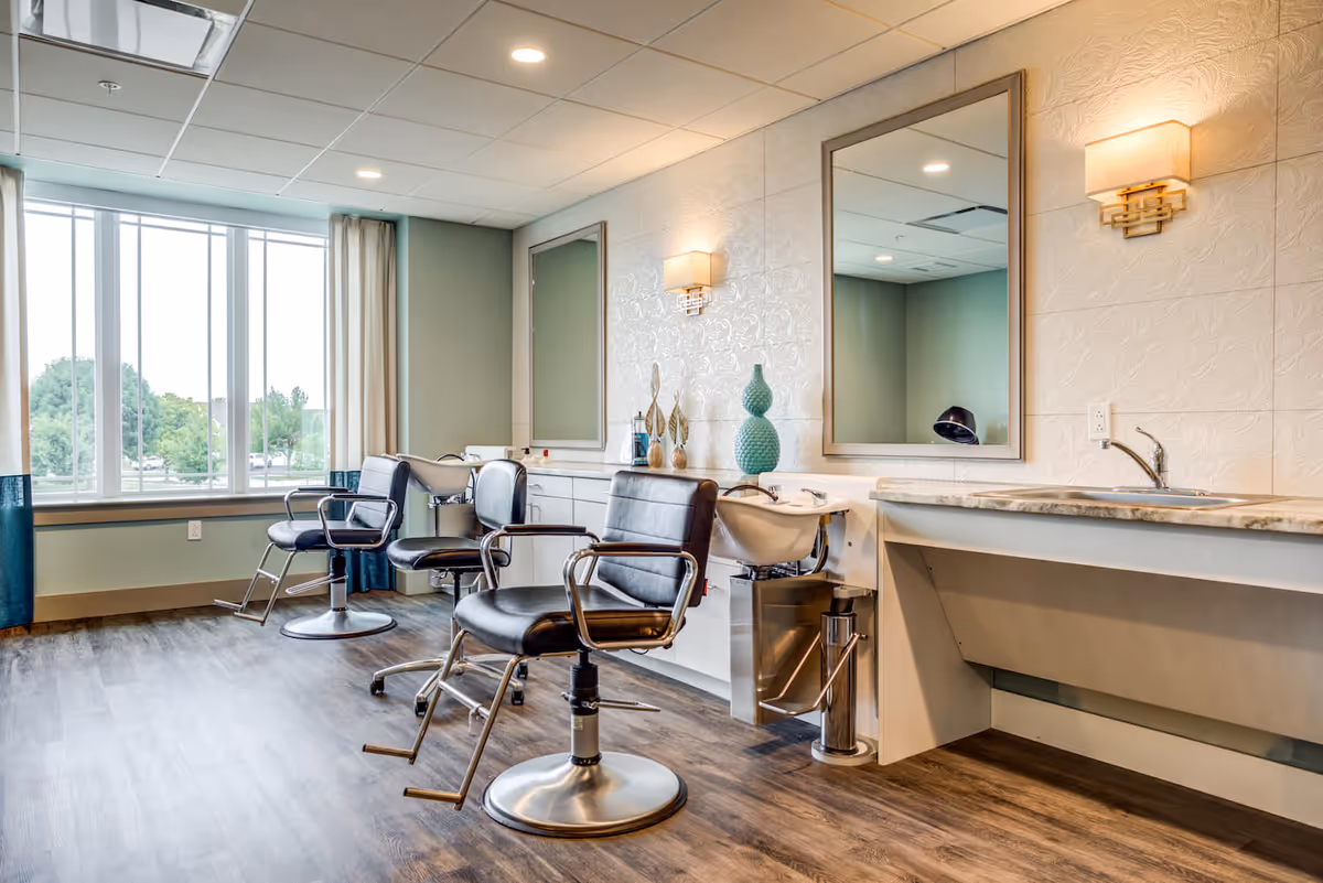 Interior of a salon area with three black salon chairs in front of two large mirrors and hair washing sinks. The room has wood flooring, light-colored walls with decorative wall sconces, and a large window with curtains letting in natural light.