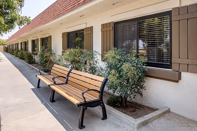 Outdoor walkway alongside a building with windows and brown shutters, featuring two wooden benches with black metal armrests and legs, and small green bushes planted in a narrow garden bed.