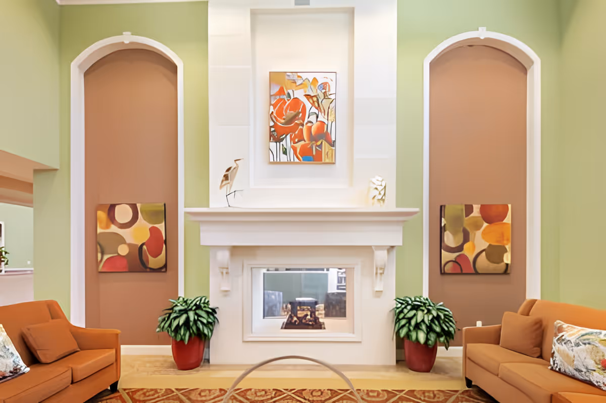 A cozy living room area with two orange sofas facing each other, a white fireplace in the center with a colorful abstract painting above it, two potted plants on either side of the fireplace, and two arched wall niches each containing an abstract painting. The walls are painted light green with brown accent panels in the niches.