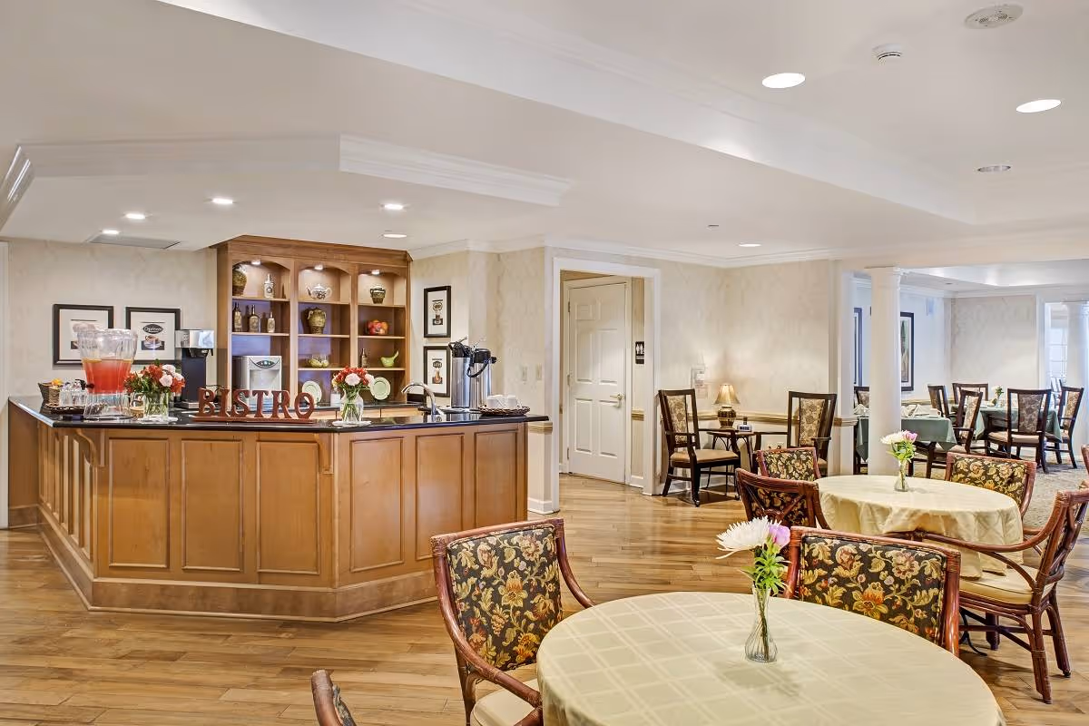 A cozy dining area in a senior living facility featuring round tables covered with light-colored tablecloths and floral centerpieces. The room has wooden flooring and floral upholstered chairs. In the background, there is a wooden bistro counter with decorative items and a beverage dispenser. The walls are light-colored with framed artwork, and the space is softly lit with ceiling lights and a small table lamp.