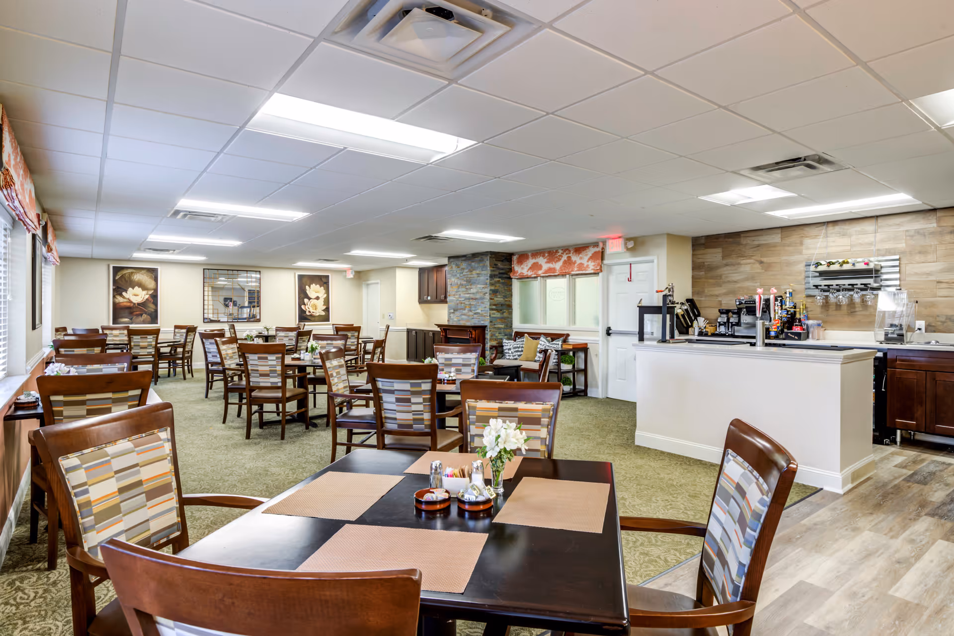 A spacious dining room in a senior living facility with multiple wooden tables and chairs arranged neatly. The room features carpeted flooring with a section of wood-like flooring near a counter area equipped with coffee machines and beverage dispensers. The walls are decorated with floral artwork and a large mirror, and there are windows with patterned valances allowing natural light to enter.