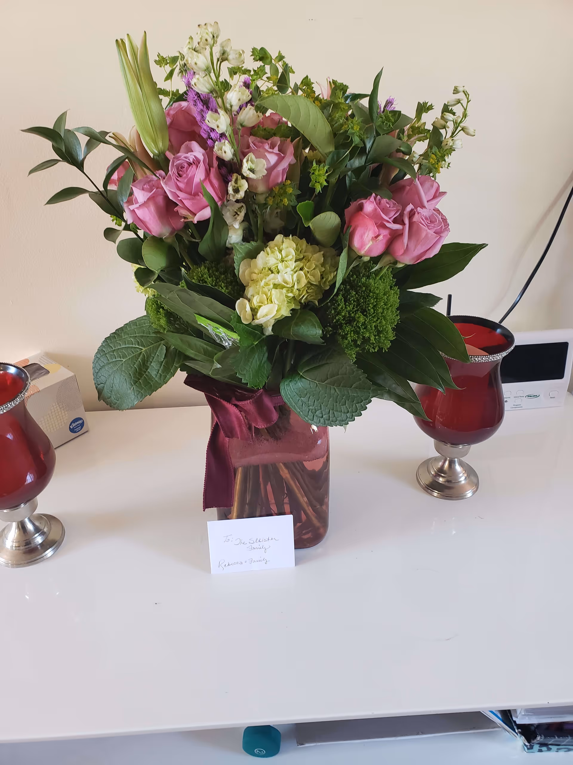 A bouquet of flowers in a glass vase with a burgundy ribbon, placed on a white surface. The bouquet includes pink roses, white flowers, green leaves, and other assorted greenery. There is a small card in front of the vase. On either side of the vase are two red candle holders with silver bases. In the background, there is a box of Kleenex tissues and a small electronic device with a screen and buttons.