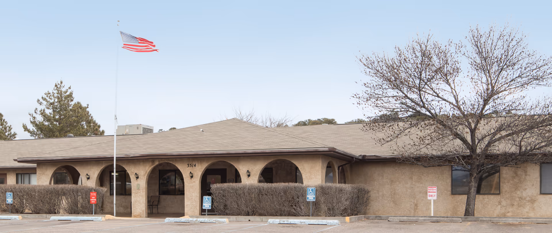 Exterior front view of a single-story building with a beige stucco facade and arched entryways. An American flag is flying on a flagpole in front of the building, and there are several parking spaces, including handicapped spots, visible in the foreground. There is a leafless tree on the right side of the building.