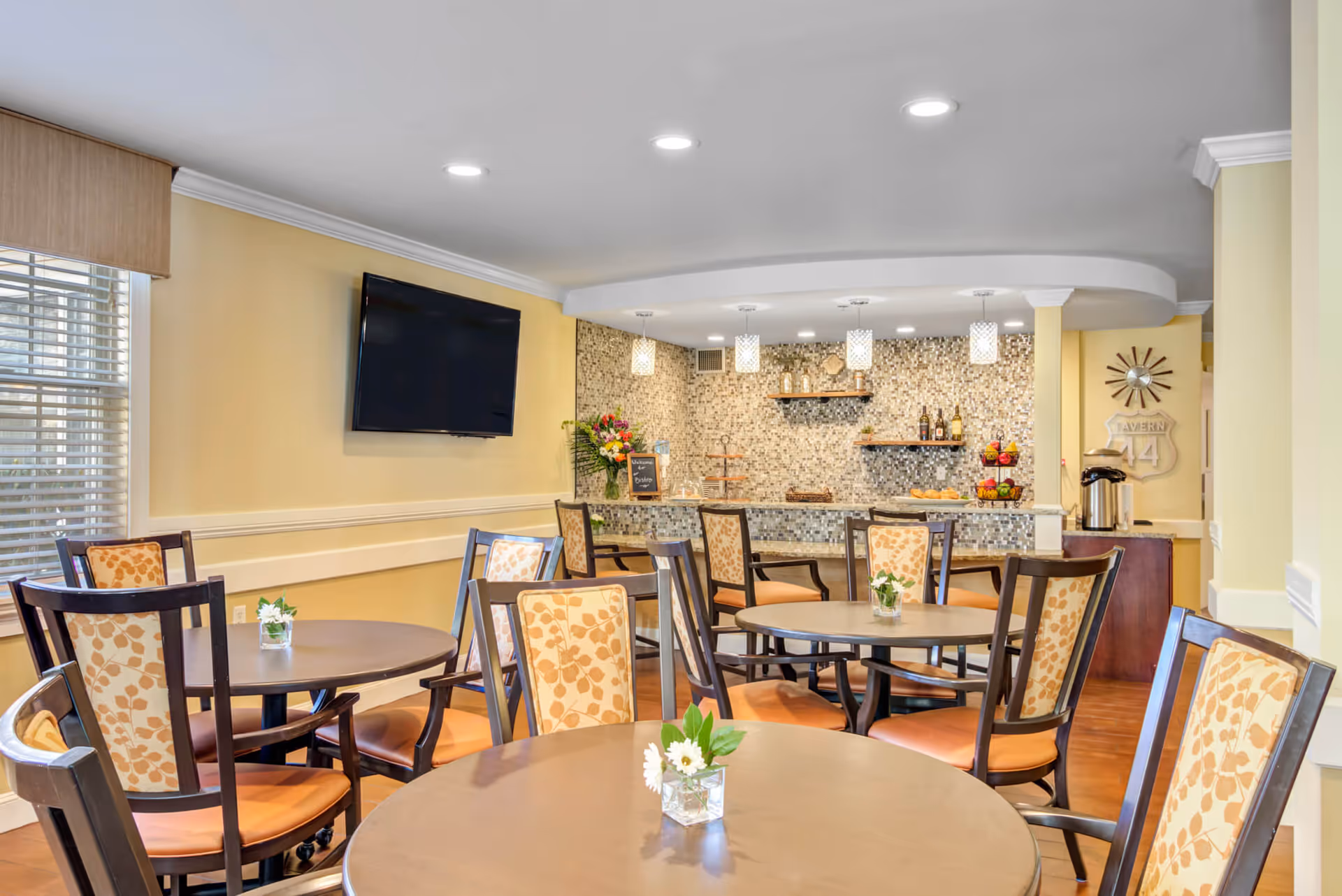 A dining area with round tables and chairs featuring floral patterned upholstery. The room has yellow walls, a mounted flat-screen TV, and a mosaic tile backsplash behind a counter with shelves holding bottles and decorative items. Small flower arrangements are placed on the tables.