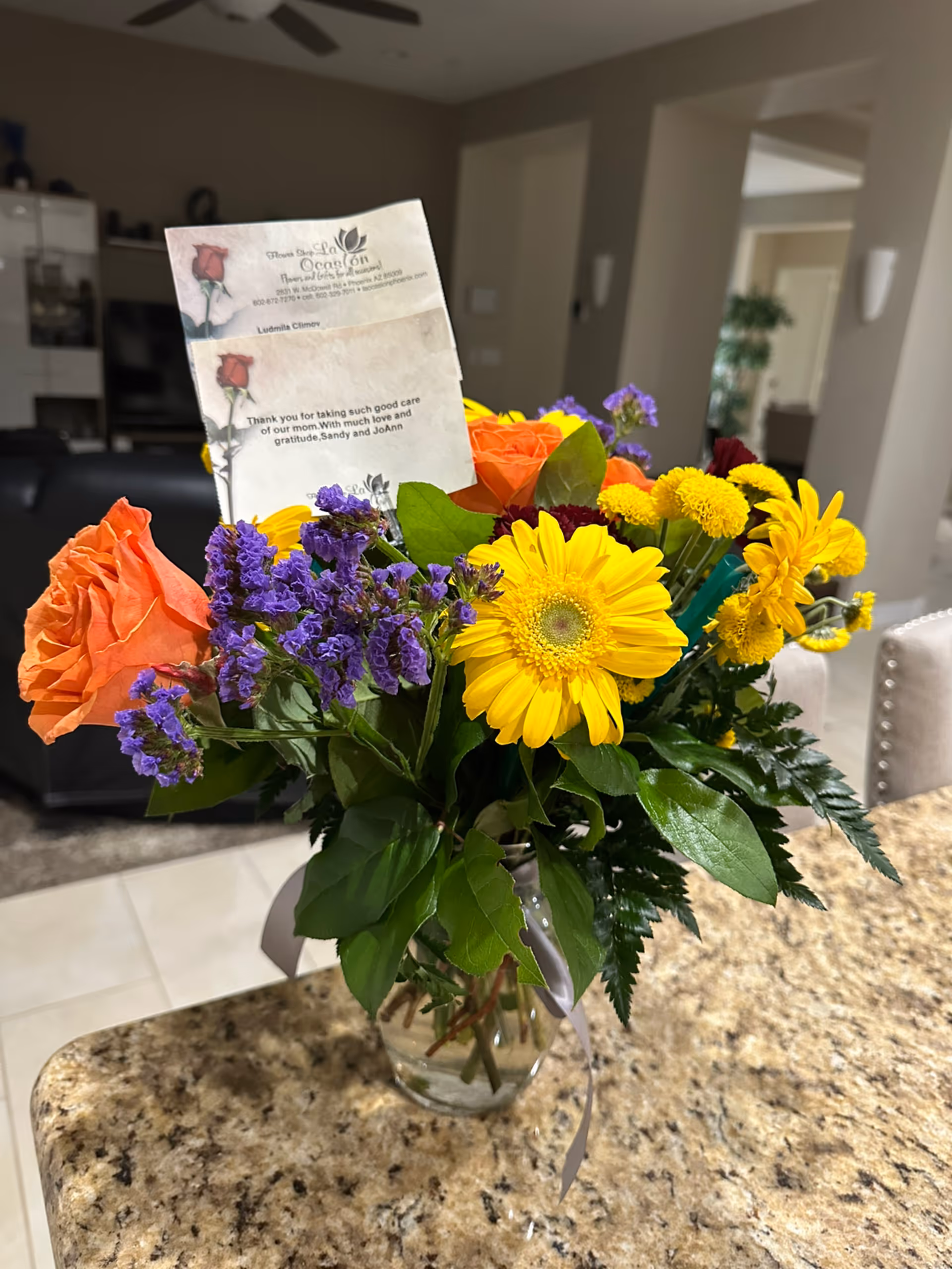 A colorful bouquet of flowers including orange roses, yellow daisies, and purple statice in a glass vase on a granite countertop. A card with a thank you message is placed among the flowers. The background shows a living area with a ceiling fan, furniture, and an open doorway.