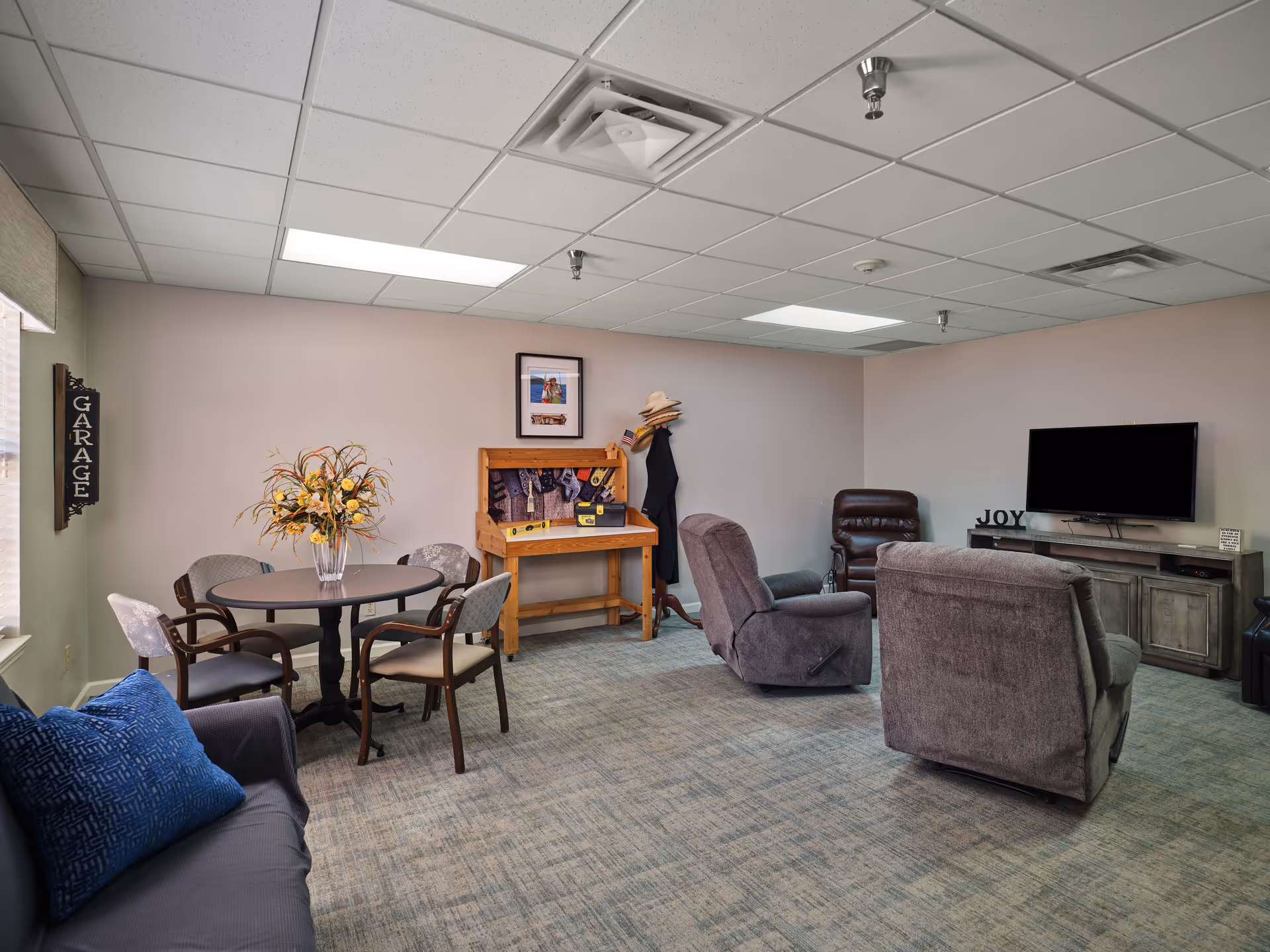 A cozy living room area with a round table and four chairs on the left, a gray couch with a blue pillow in the foreground, two recliner chairs facing a wall-mounted flat-screen TV on a wooden media console. A wooden coat rack with hats and a workbench with tools are against the back wall. The room has a carpeted floor and a drop ceiling with fluorescent lights.