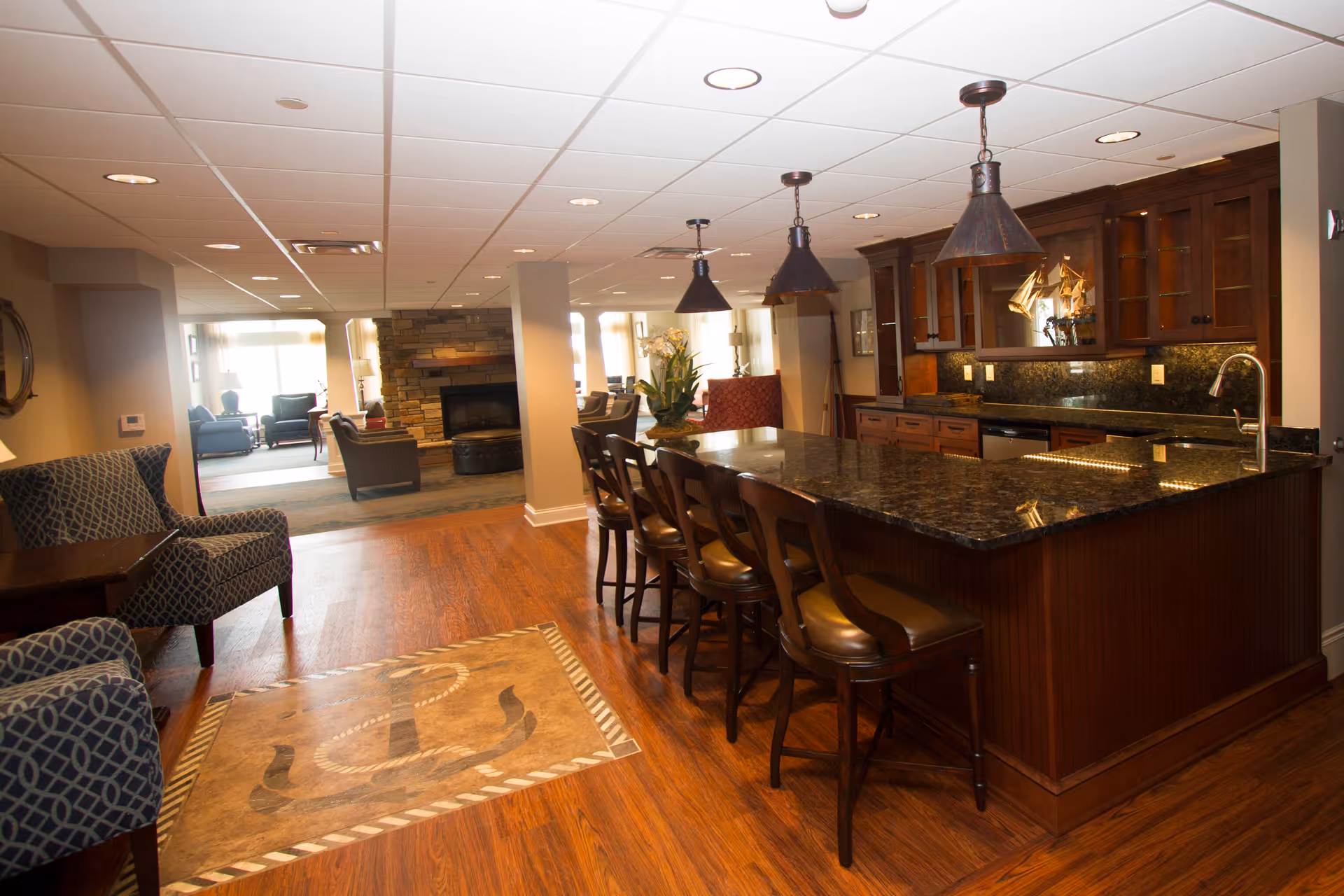 Warm communal interior with a large dark granite kitchen island and bar stools, pendant lights, hardwood floors and a seating area with a fireplace in the background.