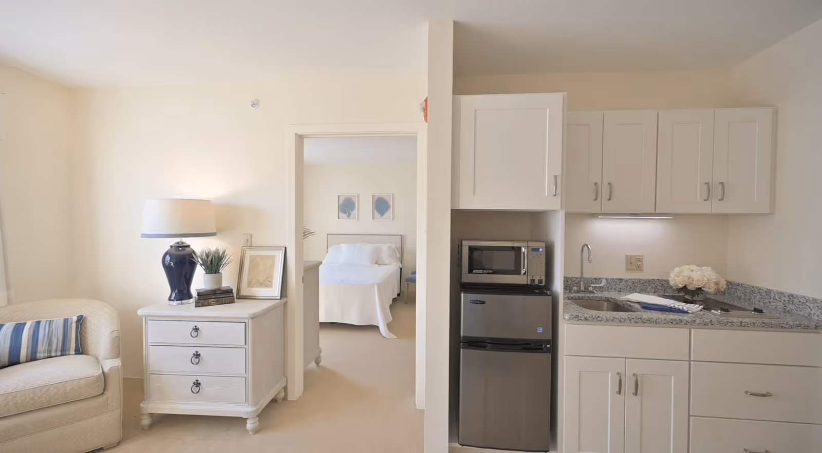 A bright and clean interior of a senior living facility room featuring a small kitchenette with white cabinets, a microwave, and a mini refrigerator. To the left, there is a cozy sitting area with a beige armchair and a white side table holding a blue lamp, a small plant, and framed pictures. In the background, a bedroom with a neatly made bed and two framed artworks on the wall is visible through an open doorway.