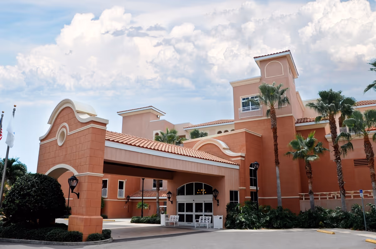 Exterior view of Westminster Shores facility showing a large building with terracotta-colored walls, a covered entrance with arches, palm trees, and a partly cloudy sky.