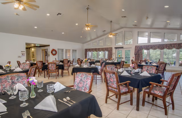 A spacious dining room with multiple tables covered in black tablecloths, each set with folded white napkins, glassware, silverware, and small flower arrangements. The room has large windows with floral curtains, ceiling fans, and tiled floors, creating a bright and welcoming atmosphere.