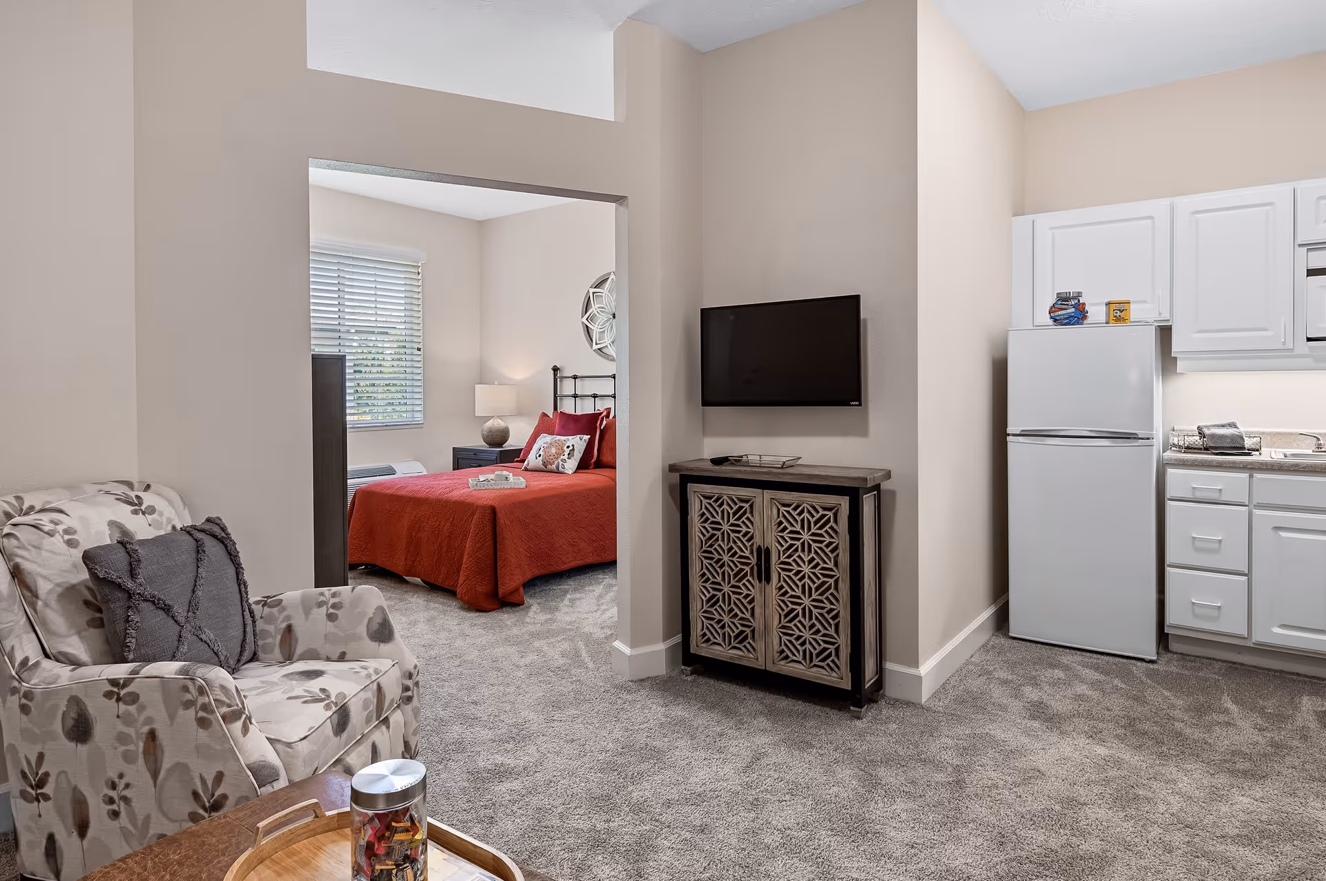 Interior view of a senior living facility apartment showing a cozy living area with a floral armchair and a small wooden table, a wall-mounted TV above a decorative cabinet, a compact kitchen with white cabinets and a refrigerator, and a bedroom with a red bedspread visible through an open doorway.