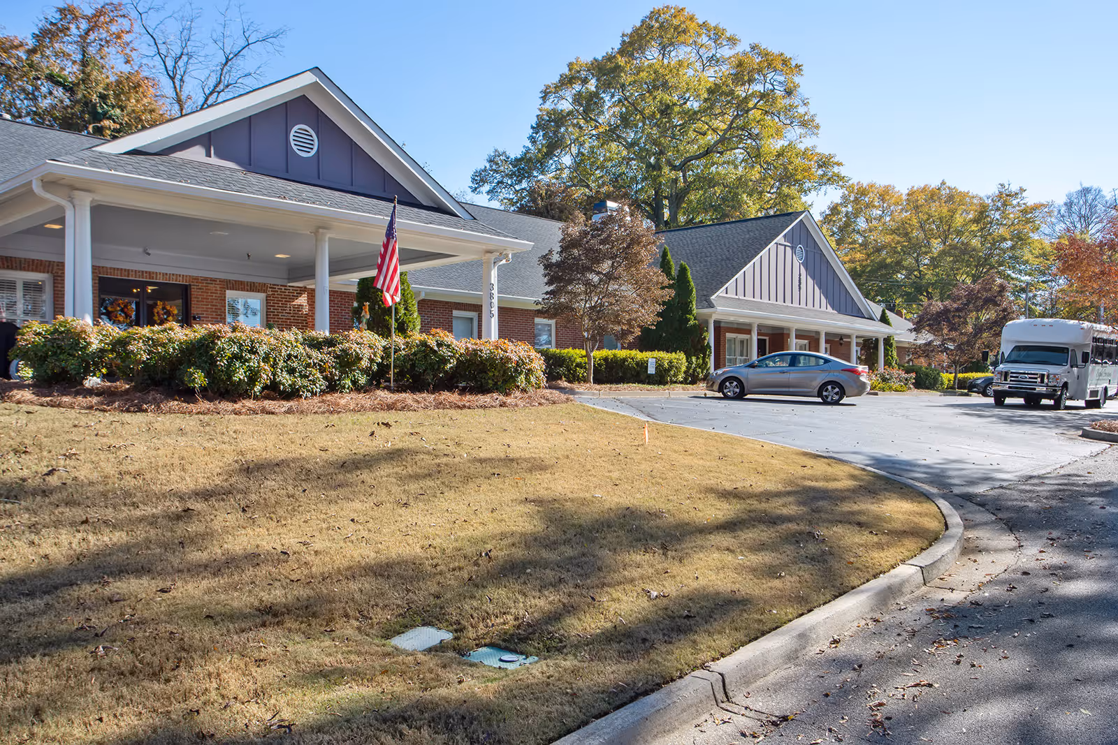 Exterior view of a single-story assisted living facility building with a covered entrance, an American flag, bushes, a parked car, and a shuttle bus in the parking lot under a clear blue sky.