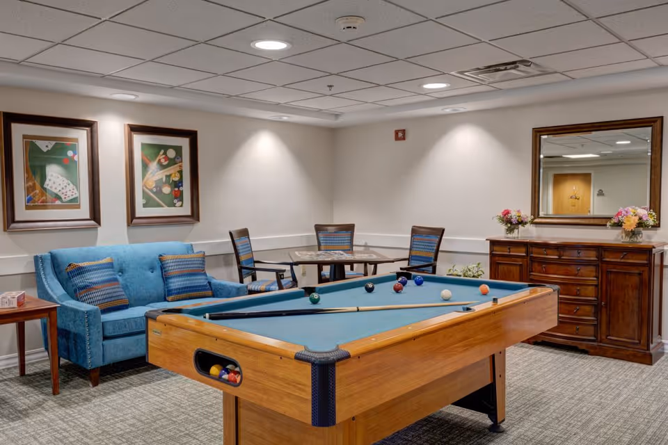 A recreational room featuring a pool table with balls and cues, a blue upholstered sofa with patterned cushions, a small table with four chairs, framed artwork on the wall, and a wooden dresser with a large mirror and flower arrangements on top.