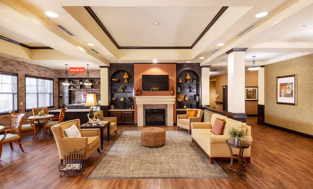 A spacious and well-lit living room area in a senior living facility featuring a beige sofa with pillows, two armchairs, a round ottoman, and a fireplace with a mounted TV above it. The room has wooden flooring, built-in dark wood shelves with decorative items, and large windows letting in natural light. There is also a small dining area with tables and chairs visible to the left.