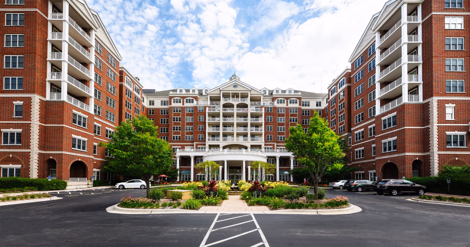 Front view of a large red-brick multi-story senior living building with a landscaped circular driveway and parked cars.
