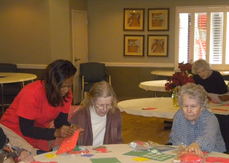 A group of elderly women sitting around a table engaged in a craft activity with paper and decorations in a well-lit room with round tables and framed artwork on the wall.