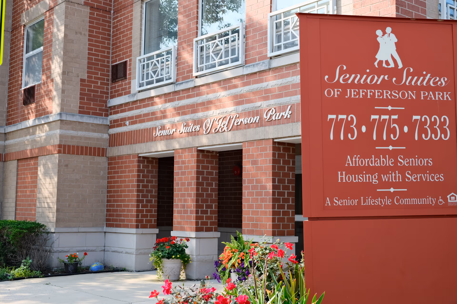 Entrance and red sign for Senior Suites of Jefferson Park next to a brick building entrance with potted flowers.